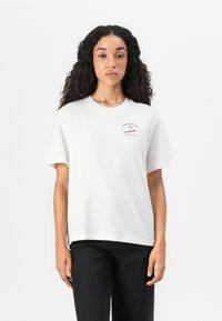 Donna con lunghi capelli ricci che indossa una maglietta bianca a maniche corte con il logo rosso "Billabong Surf Club" e pantaloni neri, in piedi contro uno sfondo bianco.