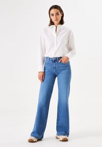 Witte button-up shirt gecombineerd met lichtblauwe flared jeans en witte sneakers met bruine zolen. Eenvoudige, klassieke stijl met een casual pasvorm.