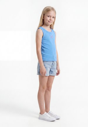 Νεαρό κορίτσι που στέκεται, φοράει μπλε αμάνικη μπλούζα με σχέδιο πεταλούδας, ανοιχτόχρωμα τζιν σορτς και λευκά αθλητικά παπούτσια σε λευκό φόντο.