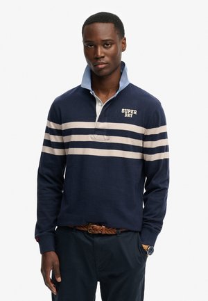 Man draagt een marineblauw rugbyshirt met witte horizontale strepen en het logo "Super Dry", gecombineerd met een donkere broek en een gevlochten bruine riem.