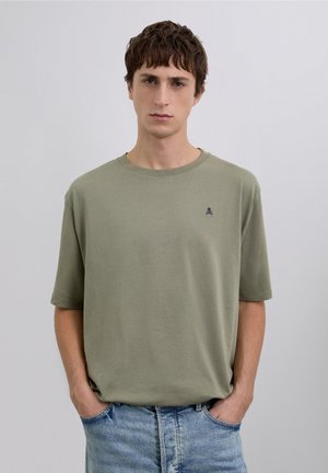 Camiseta de algodón verde oliva claro con un corte relajado, cuello redondo y mangas cortas; presenta un pequeño logo de calavera azul marino en el pecho.