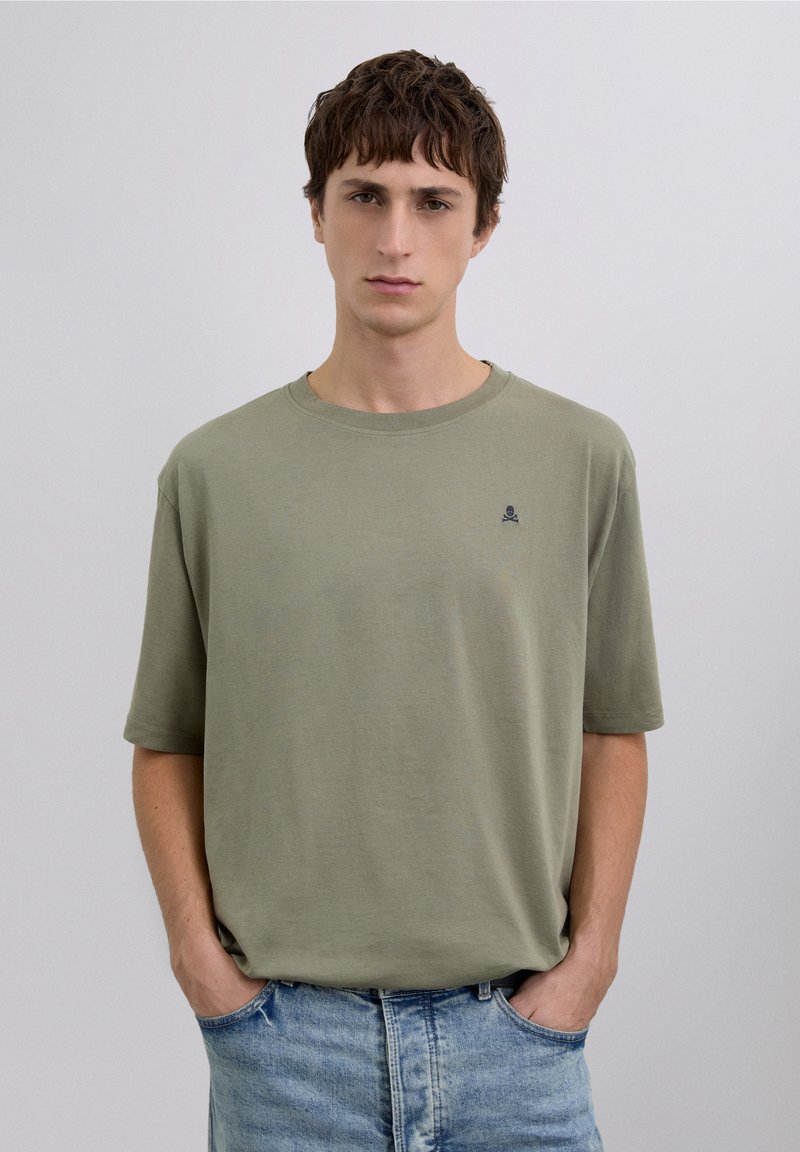 Camiseta de algodón verde oliva claro con un corte relajado, cuello redondo y mangas cortas; presenta un pequeño logo de calavera azul marino en el pecho.