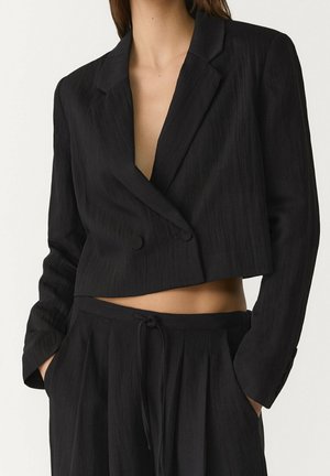 Zwarte cropped dubbelgeknoopte blazer gecombineerd met zwarte hooggetailleerde broek met trekkoord, handen in zakken, toont de buik.
