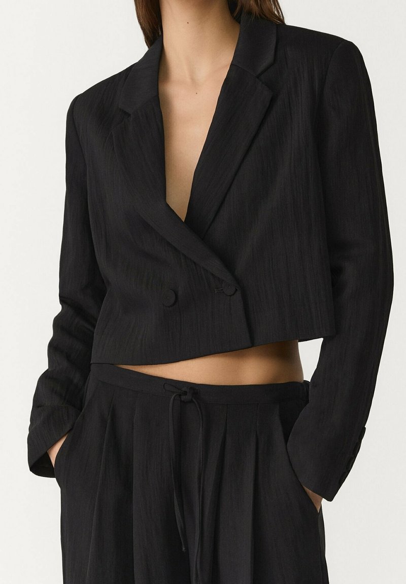 Veste croisée noire raccourcie associée à un pantalon noir taille haute avec cordon, mains dans les poches, dévoilant le ventre.