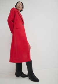 Manteau en mélange de laine rouge avec un col à revers et des poches latérales, associé à des bottes de cowboy en cuir noir à motifs. Texture lisse, design longueur genou.