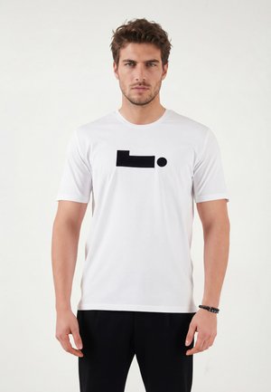 Homme aux cheveux courts et bouclés portant un t-shirt blanc avec un motif géométrique noir et un pantalon noir sur un fond blanc uni.