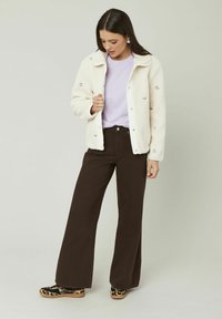 Veste blanche duveteuse avec broderies florales sur un t-shirt violet clair, associée à un pantalon marron foncé taille haute et des chaussures à motifs.