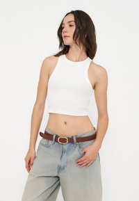 Femme portant un crop top blanc sans manches, un jean bleu clair et une ceinture marron, debout avec une main dans la poche devant un fond uni.