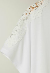 Top bianco con un dettaglio in pizzo sulla spalla e motivi floreali. Tessuto morbido, vestibilità rilassata e maniche corte.