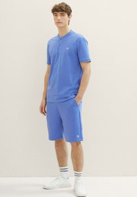 Pánská modrá polokošile s krátkými rukávy a knoflíky, v kombinaci s odpovídajícími modrými šortkami. Vyrobena z hladkého materiálu; obsahuje logo na hrudi a na šortkách.