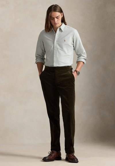 Φόρεμα ανοιχτοπράσινο πουκάμισο με κουμπιά και λεπτομέρειες πόλο, σε συνδυασμό με σκουροπράσινο παντελόνι καφέ και καφέ δερμάτινα παπούτσια. Μακριά μαλλιά, χέρια στις τσέπες.