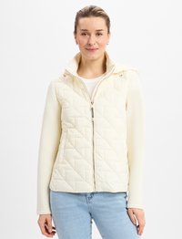 Gepolsterte cremfarbene Weste mit hohe Kragen, Reißverschluss und schlankem Design, die über einem hellen Top getragen wird. Auffälliges Rautenmuster und weiche Textur.