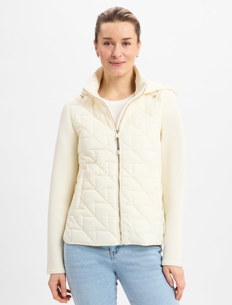 Gepolsterte cremfarbene Weste mit hohe Kragen, Reißverschluss und schlankem Design, die über einem hellen Top getragen wird. Auffälliges Rautenmuster und weiche Textur.