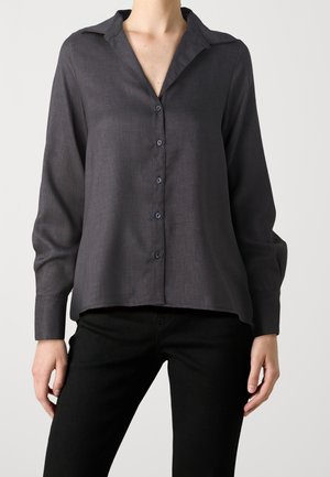 Woman wearing a dark gray long-sleeve button-up shirt with a collar, paired with black pants, standing against a plain background.