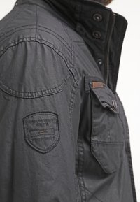 Gros plan sur l'épaule d'une veste gris foncé avec un patch cousu indiquant "Vintage Travel Route Team Spirit" et une poche poitrine boutonnée.