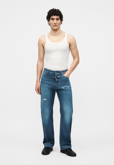 Άνδρας που στέκεται με το ένα χέρι στην τσέπη, φοράει λευκό φανελάκι, μπλε τζιν με ελαφρές φθορές και μαύρα παπούτσια σε απλό φωτεινό φόντο.