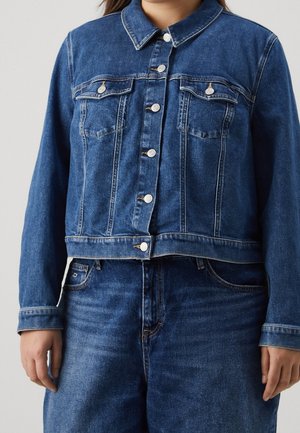Kvinde iført en blå denimjakke med forreste knapper og brystlommer kombineret med blå denimjeans.
