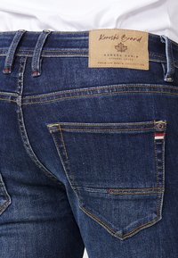 Jeans de mezclilla oscura con un parche de logo de cuero, costura visible, dos bolsillos traseros con acentos de color y una ligera textura desvanecida.