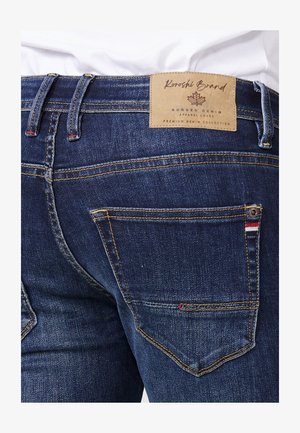 Tumedat denim teksad, millel on nahkne logo pealt, nähtav õmblus, kaks tagakotti koos värvilise aktsendiga ja kergelt fade-efekt.