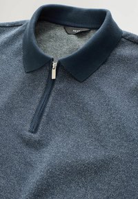Tummansininen vetoketjullinen polo-paita, jossa on ribbireunus ja teksturoitu kangas, jossa on hieno viisto raidoituskuvio. Brändilappu näkyy sisällä.