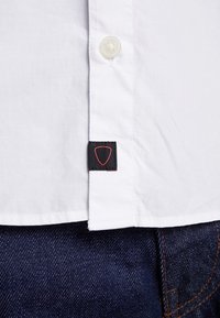 Biała koszula z krótkim rękawem z guzikiem i czarną metką, na której znajduje się czerwone, trójkątne logo, nałożona na ciemnoniebieski materiał dżinsowy.