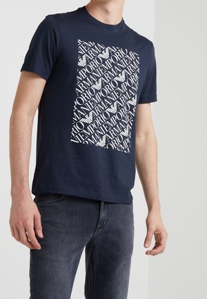 Mann trägt ein dunkelblaues Emporio Armani T-Shirt mit weißem Logo-Textmuster und dunklen Jeans vor einem einfarbigen hellen Hintergrund.