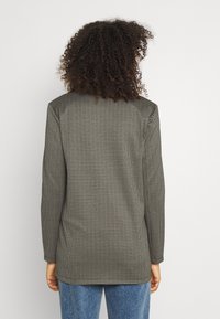 Šedý kostkovaný blejzr s dlouhými rukávy, texturovanou tkaninou a rovný střih. Má mírně delší zadní část a jemné vzory.