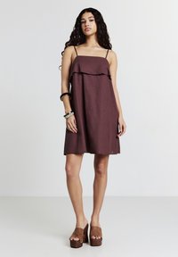 Junge Frau mit langen lockigen Haaren, die ein ärmelloses braunes Kleid, Plateau-Sandalen und Armbänder trägt und vor einem schlichten weißen Hintergrund steht.