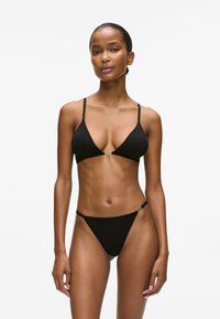 Juodas bikini komplektas, turintis trikampį viršų su reguliuojamomis petnešėlėmis ir metaliniu akcentu, derinamas su minimaliai dengiančiais dugnais. Minkšta audinio tekstūra.