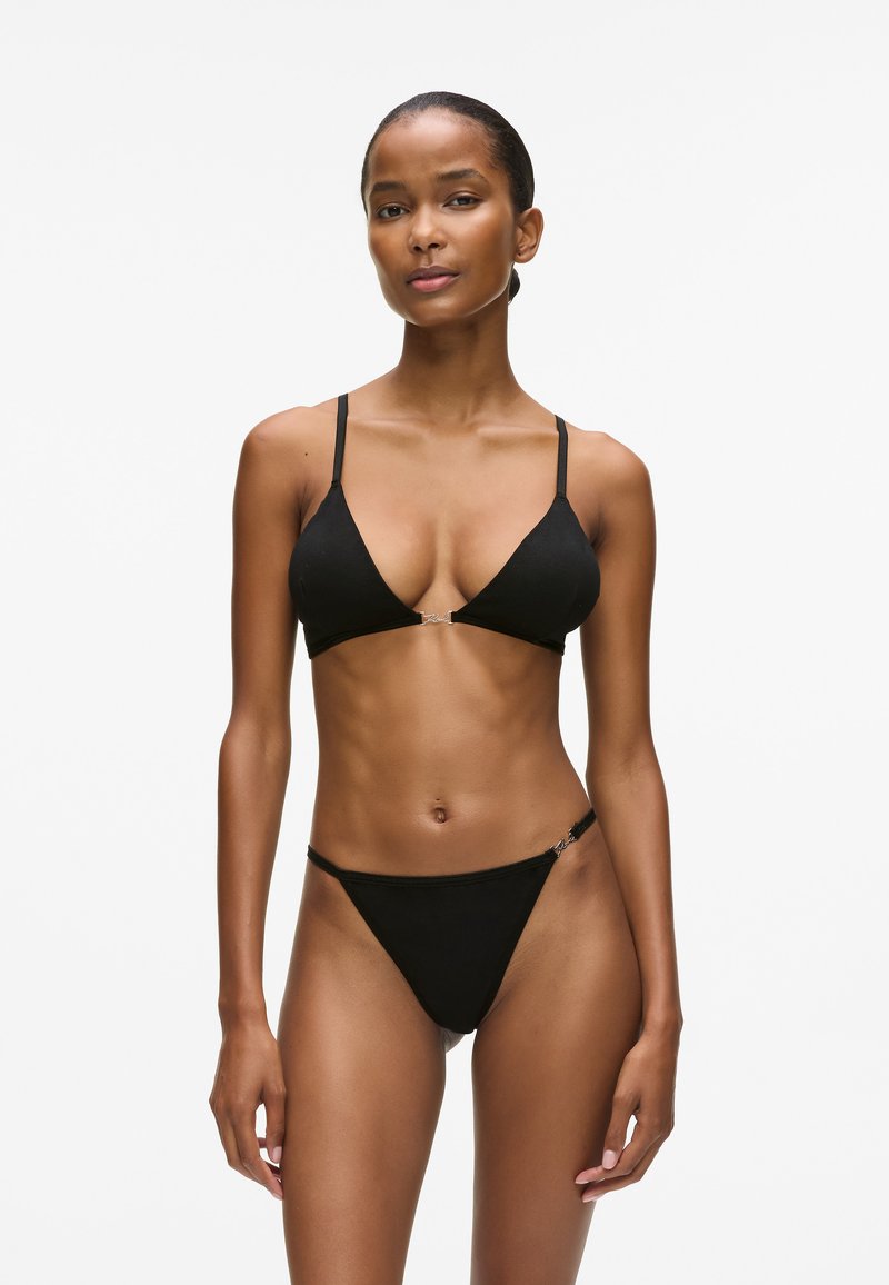 Juodas bikini komplektas, turintis trikampį viršų su reguliuojamomis petnešėlėmis ir metaliniu akcentu, derinamas su minimaliai dengiančiais dugnais. Minkšta audinio tekstūra.