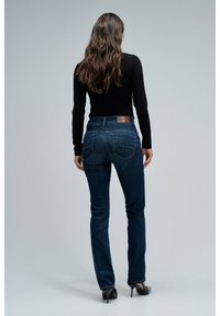Vaqueros de mezclilla azul oscuro con un diseño ajustado, que cuentan con bolsillos traseros y una etiqueta de cuero en la cinturilla. Combinados con una camiseta negra de manga larga.
