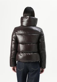 Persoon in een glanzende zwarte pufferjack met een hoge kraag, gecombineerd met donkerblauwe jeans, met de rug naar de camera tegen een effen achtergrond.