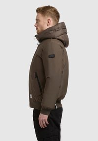 Veste matelassée marron avec capuche amovible, poches latérales zippées et poignets côtelés. Texture lisse et détails de marque minimalistes sur la manche.