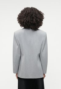 Harmaa, muotoonommeltu bleiseri, jossa on jämäkkä istuvuus, yksittäinen takatuuletus ja pidentyneet hihat. Teksturoitu kangas, jossa on sileä pinta.