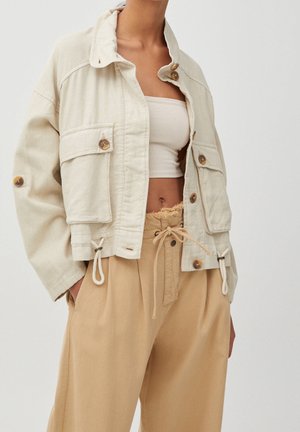 Mujer con chaqueta corta beige con grandes botones, pantalones de tiro alto con cordón color topo y top corto blanco de canalé contra fondo liso.