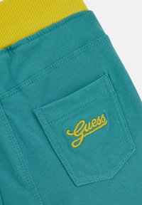 Szare dresowe spodnie z tkaniny mają jasnoniebieską, prążkowaną talię i tylną kieszeń ozdobioną żółtym haftem z logiem "Guess".