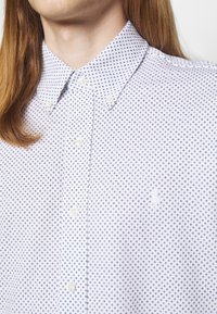 Osoba ve bílém košili na knoflíky s modrým diamantovým vzorem a dlouhými rovnými vlasy, límeček zapnutý.