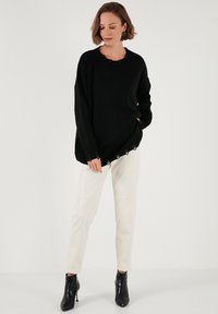 Pulover negru din tricot cu guler rotund și margine ondulată, cu detalii decupate pe mâneci. Asortat cu pantaloni crem și pantofi cu toc negri.
