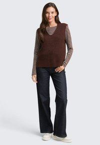 Bruine gebreide vest met een V-hals, gelaagd over een gestreept lange mouwen shirt. Draagt het met donkere wijde jeans en witte sneakers.