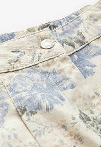 Kreemitaustaga lilleline denimpatch, millel on sinised ja rohelised lillemustrid ning hõbedane nööpdetail ülaosas.