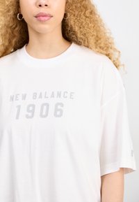 T-shirt en coton blanc présentant "NEW BALANCE 1906" en impression gris clair. Coupe décontractée avec des manches courtes et un col rond.