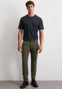 Schwarzes T-Shirt mit kurzen Ärmeln kombiniert mit olivgrünen Slim-Fit-Hosen und schwarzen Schnürstiefeln, vor einem hellen strukturierten Hintergrund.