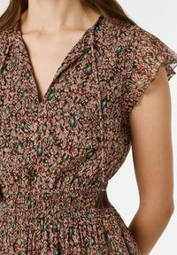 Lähipilt naisest, kes kannab pruuni lillemustriga kleiti laineliste varrukate, V-kaeluse ja kaelakaunistuseks tuttidega nööriga.