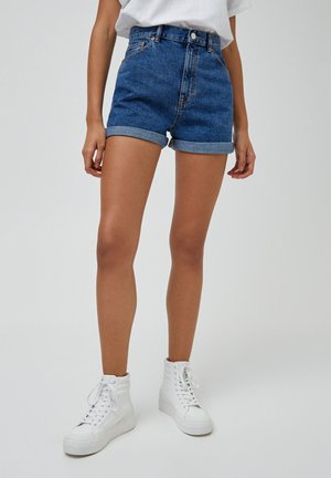 Kvinde iført op-rullede denimshorts, hvide høje sneakers og en hvid t-shirt, står på en ensfarvet baggrund.