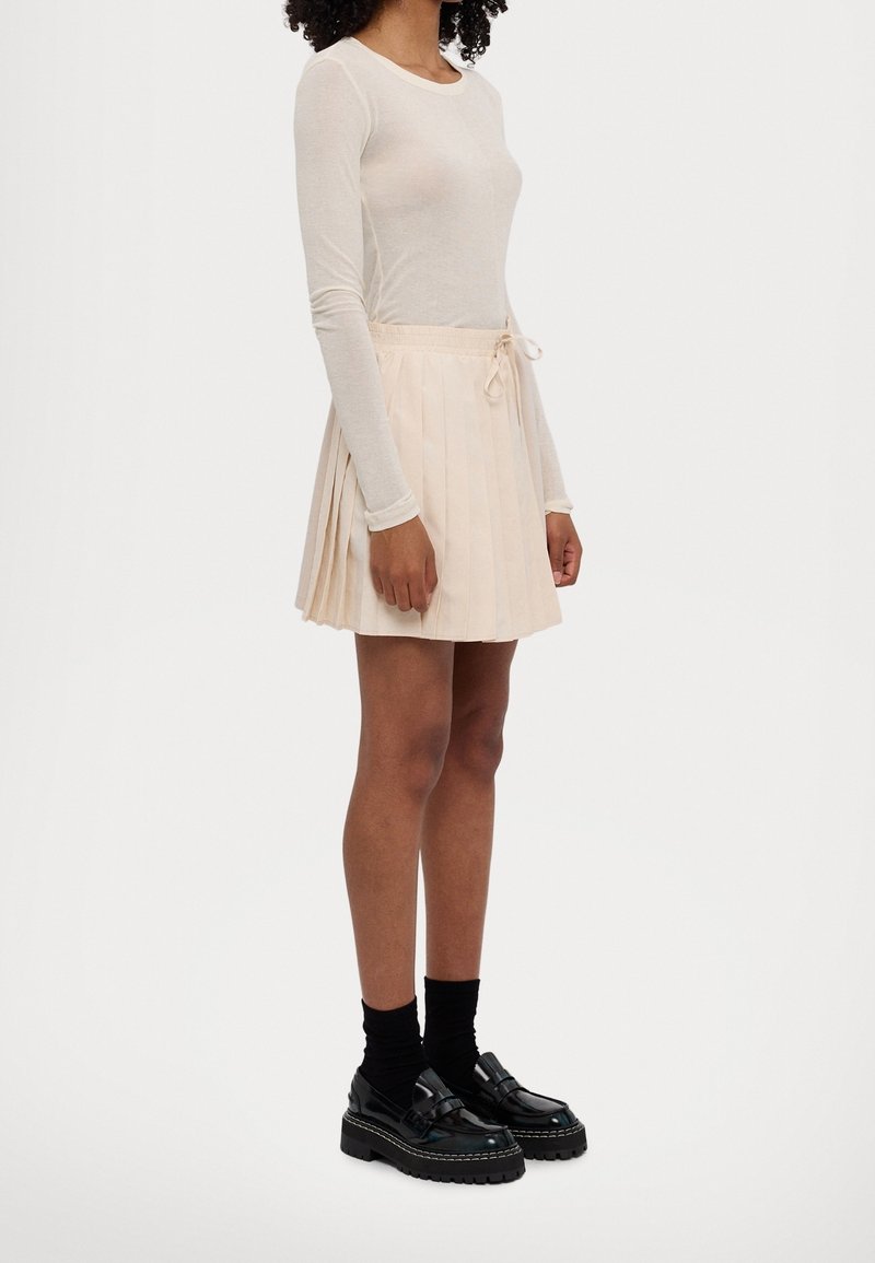 Femme portant un haut transparent à manches longues, une mini-jupe plissée beige avec taille à cordon, des chaussettes noires et des mocassins noirs épais, se tenant de côté.