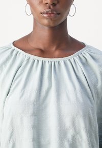 Lys blå teksturert topp med samla halslinning. Stoffet har subtile mønstre og myk fall. Modellen har på seg sølvfargede hoops.