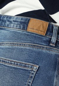 Jeans de ganga em azul médio com um rótulo de couro texturizado ostentando "American Eagle." Costuras visíveis e um design de corte reto.
