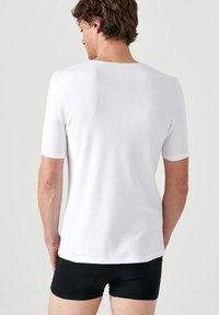 T-shirt bianco a maniche corte con colletto rotondo, texture liscia, design aderente, mostrato da dietro con pantaloni corti neri a contrasto.