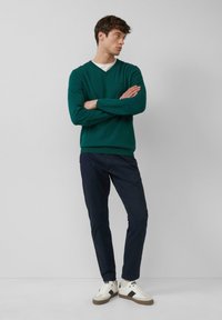 Grüner V-Ausschnitt-Pullover, marineblaue Hose und weiße Sneakers. Der Pullover hat lange Ärmel; die Hose ist schmal geschnitten. Lässiges Outfit in neutralen Tönen.