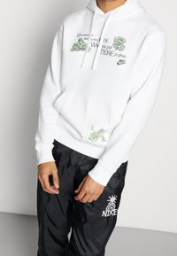 Vit hoodie med grafisk text och gröna blommönster på framsidan samt en grön djurillustration nära fickan. Tillverkad av bomull.